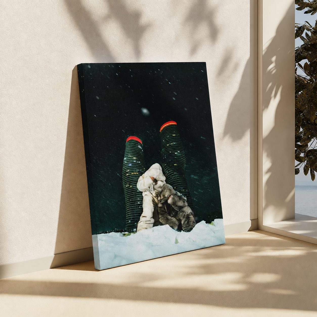 A vertical canvas art print leaning against a beige wall on a windowsill, showing a surreal photo of a person in a white coat kneeling in snow in front of two tall dark skyscrapers with red roofs under a starry sky.