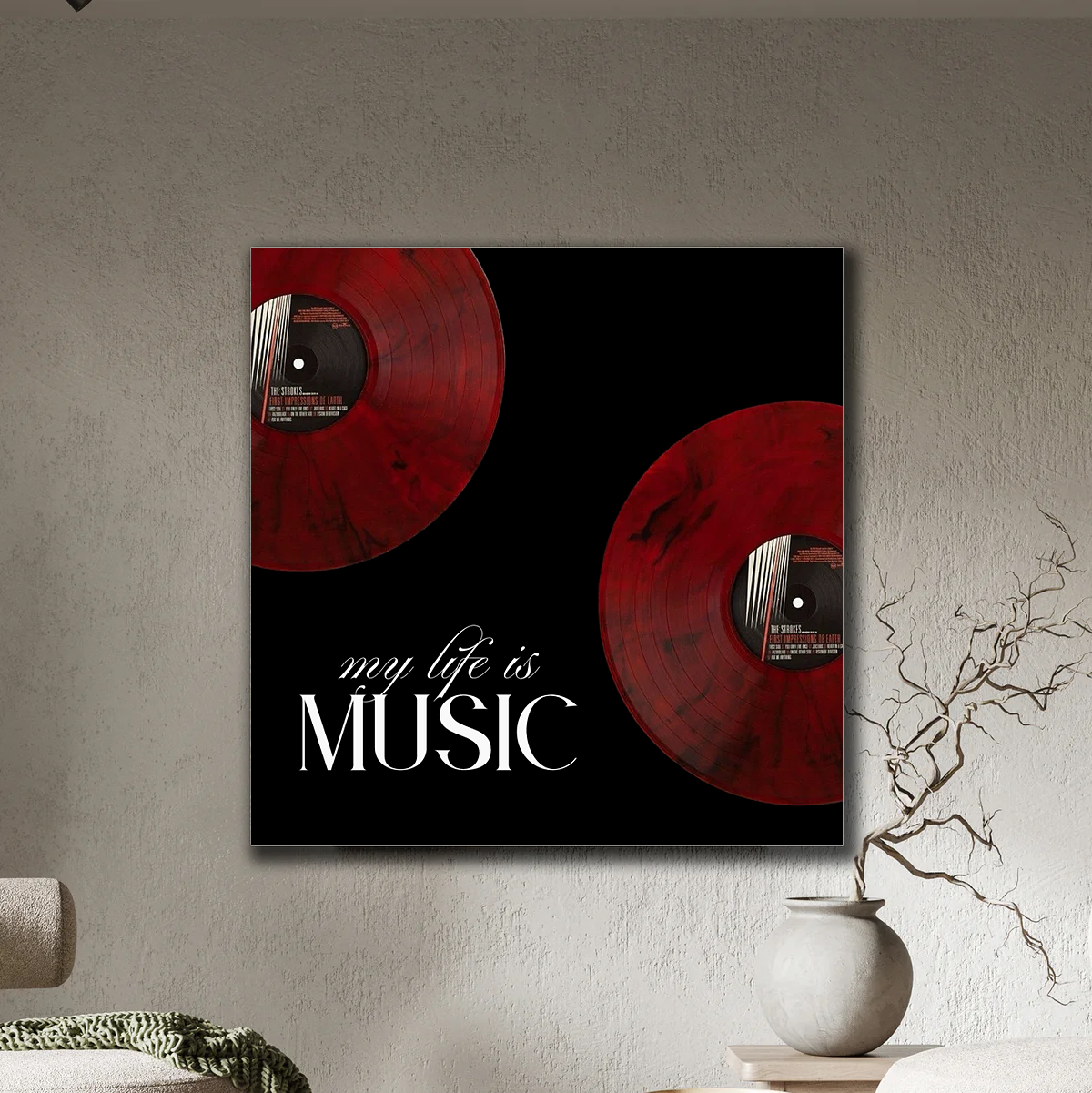 A square wall art print hanging on a textured beige wall, featuring two red vinyl records on a black background with the white text 'my life is MUSIC'. A small white vase with bare branches sits beside it, and part of a sofa is visible at the lower left.