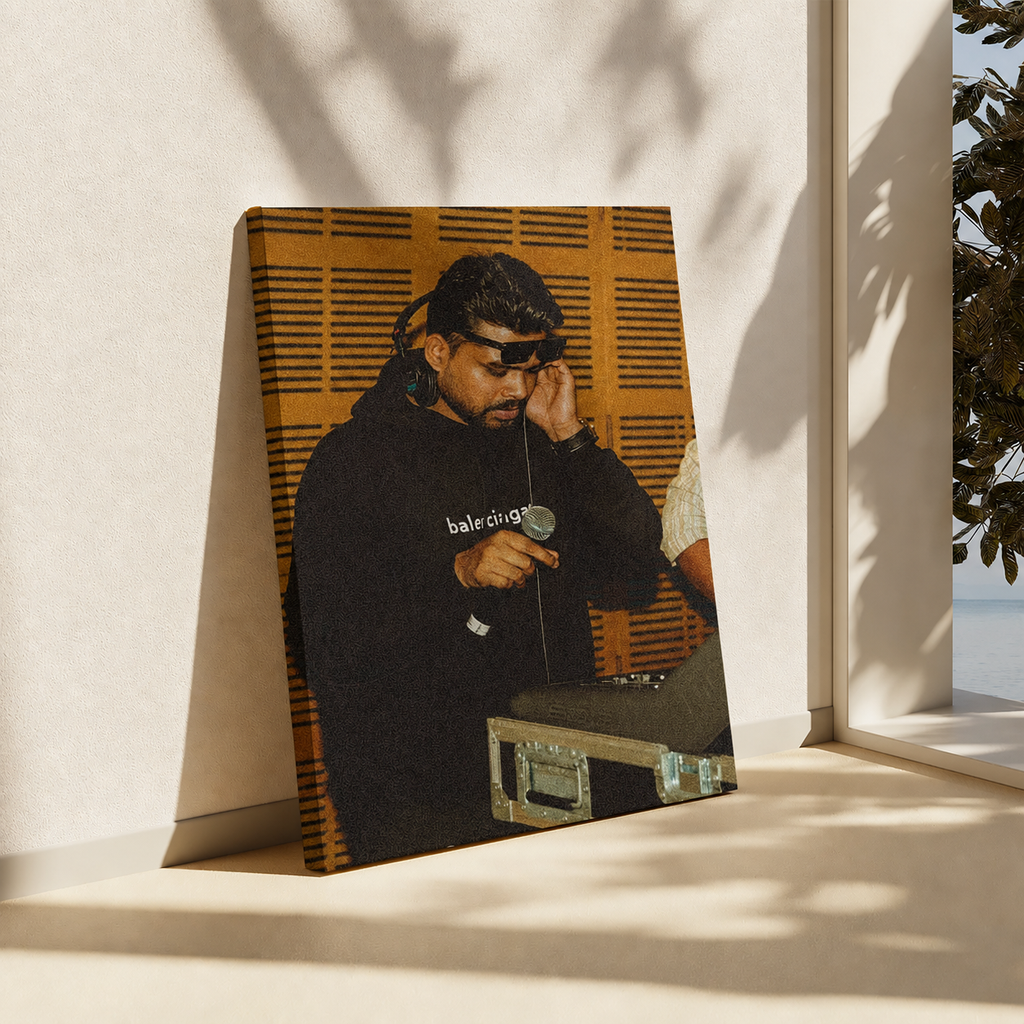 A vertical canvas print leaning against a beige wall on a windowsill, showing a photo of a man in a black hoodie with 'baleo' text. The print features a warm orange-brown slatted background and is framed by soft window shadows.
