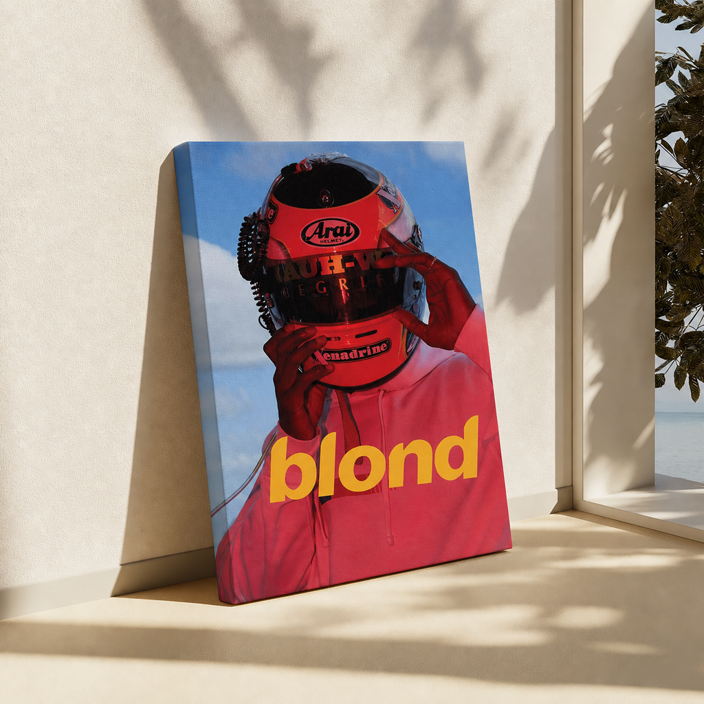 A rectangular wall art print or poster leaning against a beige wall on a windowsill, showing a person in a red racing helmet and matching red top against a blue sky. The word 'blond' appears in large yellow letters near the bottom.
