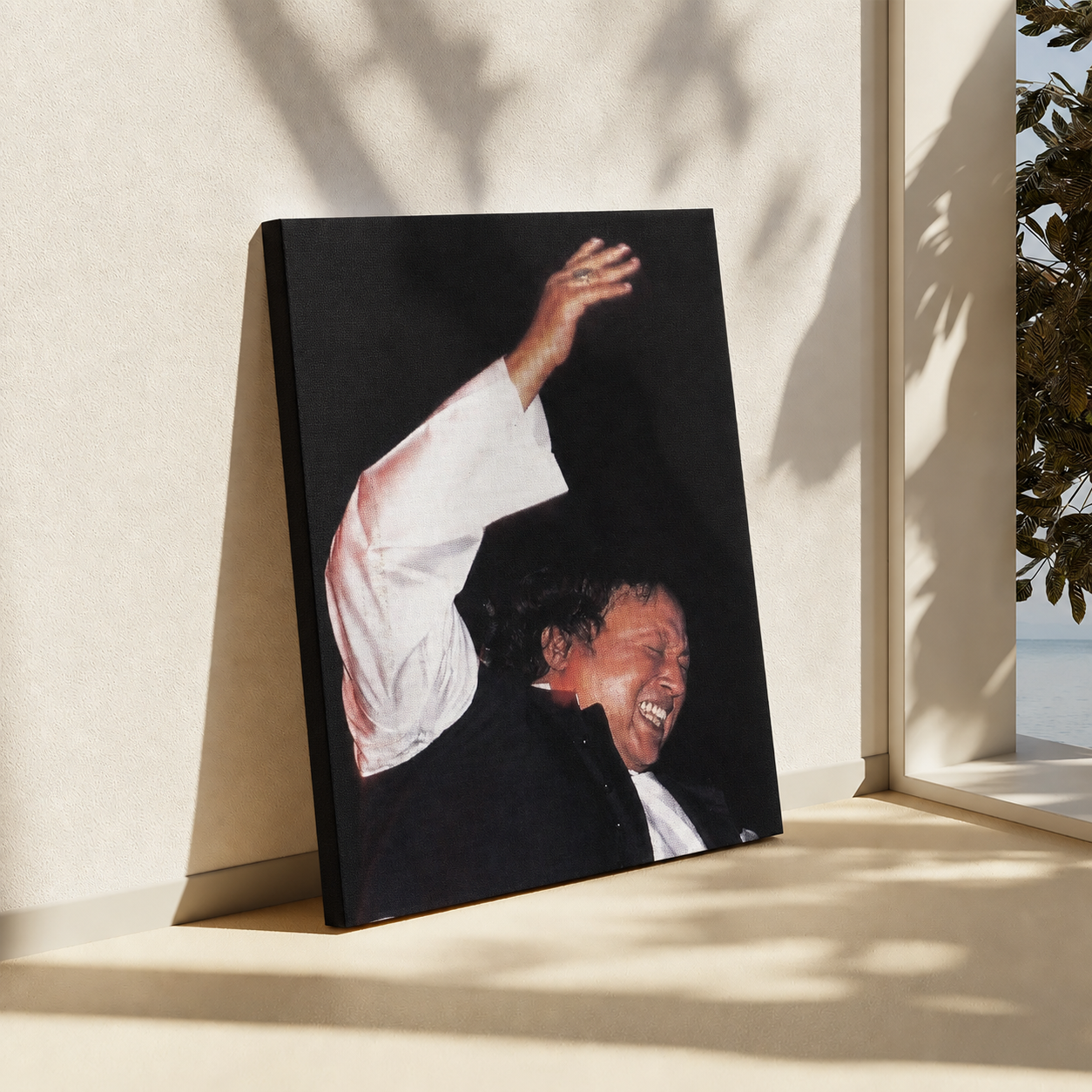 A vertical canvas print leaning against a beige wall in a sunlit interior. The artwork shows a smiling man in a black jacket and white shirt raising his arm.