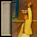 A framed art print featuring a portrait of a man in a light yellow blazer raising his arm, set against a brick wall background, with a vertical textured gray panel on the left and a black border around the frame.