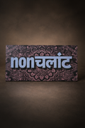 A rectangular decorative plaque or sign with the word 'nonchalant' in large white Devanagari letters over a dark paisley pattern, photographed against a dark brown gradient background.