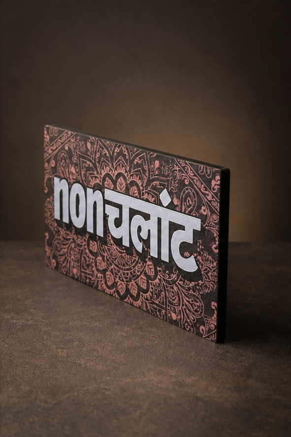 A rectangular decorative sign with the Hindi word 'nonchalant' in bold white Devanagari script over a dark brown and dusty rose paisley pattern background, standing on a dark textured surface.