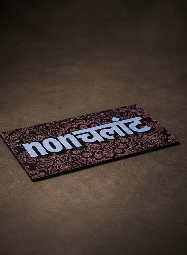 A rectangular printed card or label with the word 'non' in large white Devanagari-style lettering over a dark ornamental pattern, placed on a brown textured surface.