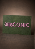 A rectangular green sign or card with the word 'AIDCONIC' in pink Devanagari-style lettering, photographed against a dark brown gradient background.