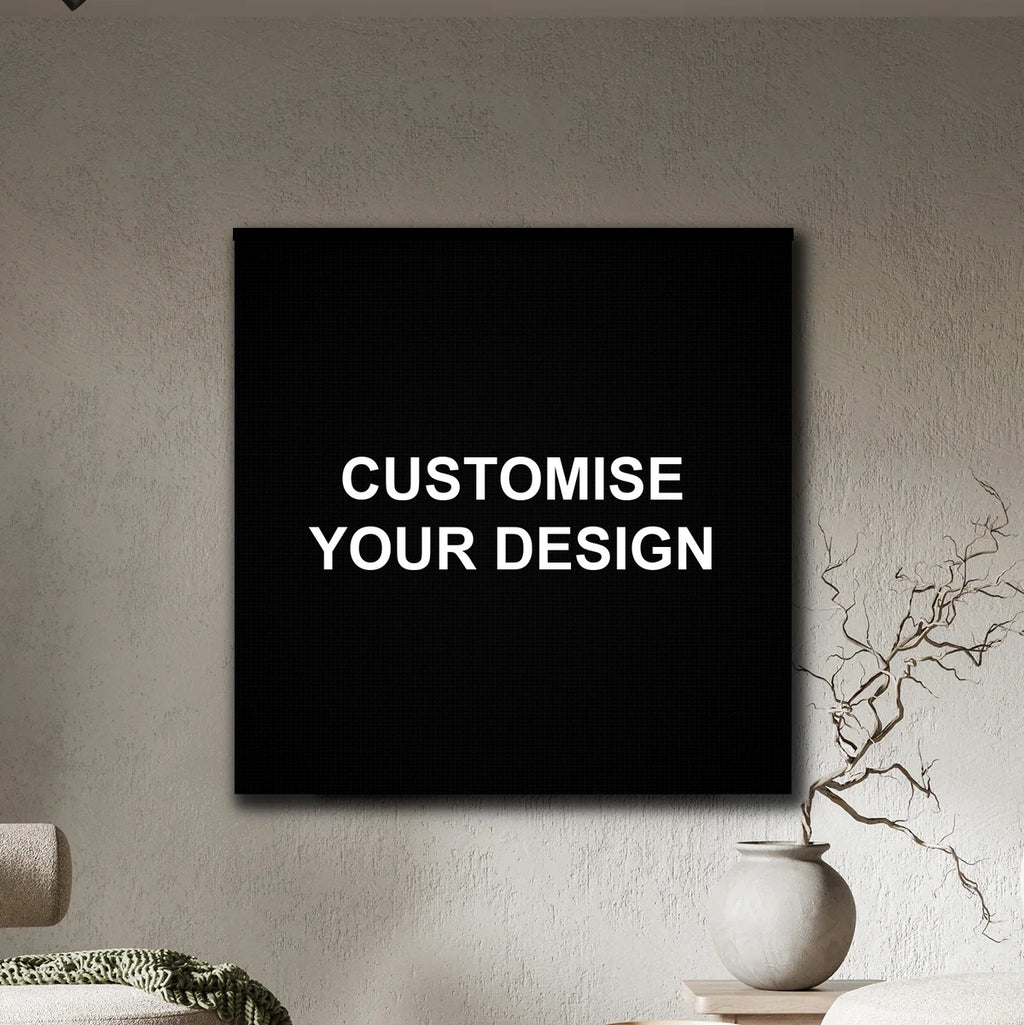 A black square wall art print or canvas hanging on a textured beige wall, with the words 'CUSTOMISE YOUR DESIGN' in white centered on it. Part of a sofa and a small vase with dried branches are visible in the room setting.