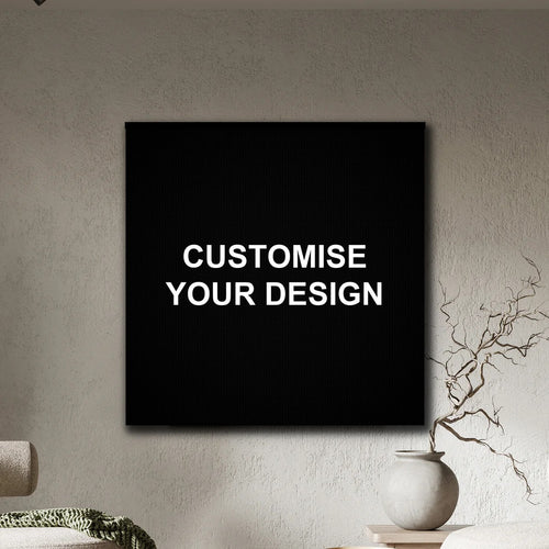 A black square wall art print or canvas hanging on a textured beige wall, with the words 'CUSTOMISE YOUR DESIGN' in white centered on it. Part of a sofa and a small vase with dried branches are visible in the room setting.