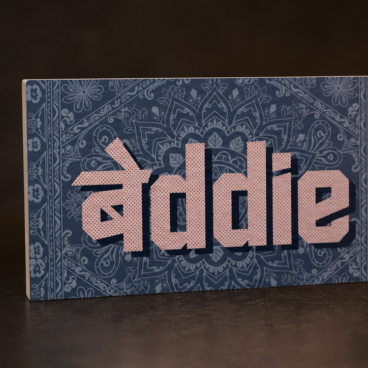 A rectangular decorative sign with the name 'Baddie' in large pink letters over a blue patterned background, photographed on a dark surface against a dark backdrop.
