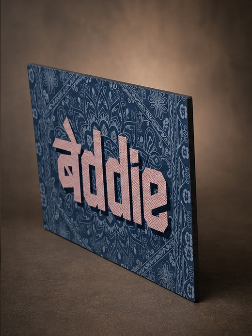 A rectangular decorative sign or canvas with a blue paisley pattern and large pink text reading 'addie', shown standing at an angle against a warm brown studio background.