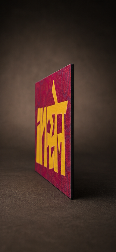 A rectangular art print or card with a textured magenta-pink background and bold yellow Devanagari lettering, shown standing upright on a dark brown surface against a dark brown backdrop.