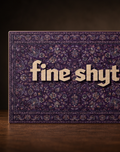 A rectangular product with a dark purple ornate patterned surface and large beige text reading 'fine shyt' across the front, photographed on a wooden surface against a dark brown background.